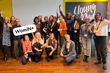 Team photo at the cinfo Forum 2024 Group of women posing indoors waving at camera, sign reads 'WomIN+'.